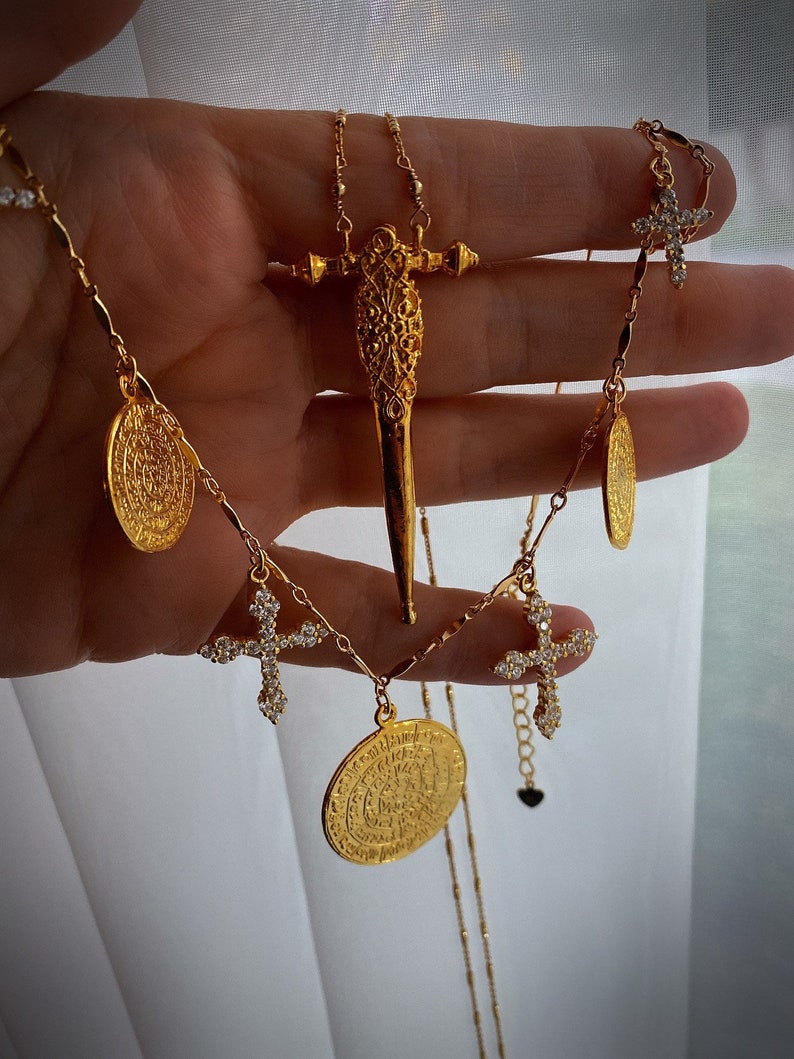 Puede incluir: Un collar de tono dorado que presenta un colgante central en forma de espada con detalles de filigrana, acentuado por m&uacute;ltiples dijes redondos estilo moneda y peque&ntilde;os colgantes de cruz incrustados con piedras claras brillantes.