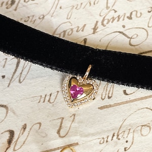 Op de afbeelding: Een zwarte fluwelen choker ketting met een gouden hartvormige hanger. De hanger is bezet met kleine heldere steentjes rondom en een roze hartvormige steen in het midden. De achtergrond is crèmekleurig papier met handschrift.