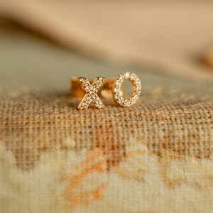 May include: A gold-colored ring with the letters "XO" encrusted with small, clear gemstones. The ring is set against a textured, tan-colored surface, creating a contrast between the shiny metal and the neutral background. The ring is a piece of jewelry.