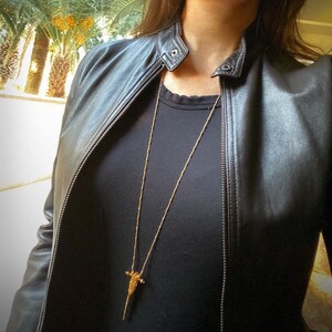 Puede incluir: Un collar largo de tono dorado con un colgante delgado y texturizado en forma de punta o daga. El collar se lleva sobre una camiseta negra lisa y una chaqueta de cuero negra con cremallera frontal y detalle de bot&oacute;n a presi&oacute;n en el cuello.