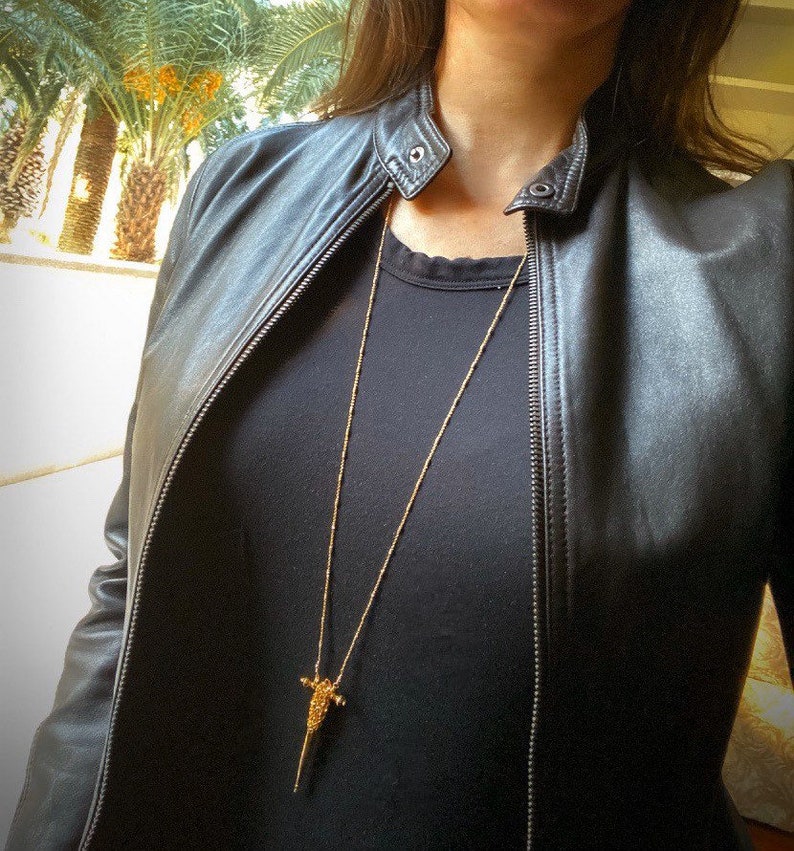Puede incluir: Un collar largo de tono dorado con un colgante delgado y texturizado en forma de punta o daga. El collar se lleva sobre una camiseta negra lisa y una chaqueta de cuero negra con cremallera frontal y detalle de bot&oacute;n a presi&oacute;n en el cuello.