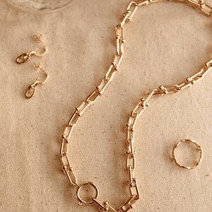 K&ouml;nnte beinhalten: Ein goldfarbenes Schmuckset mit einer klobigen Kette mit gro&szlig;em Verschluss, passenden Creolen und einem strukturierten Ring. Die Teile sind auf einer neutralen Oberfl&auml;che angeordnet und erzeugen eine zusammenh&auml;ngende und elegante &Auml;sthetik.