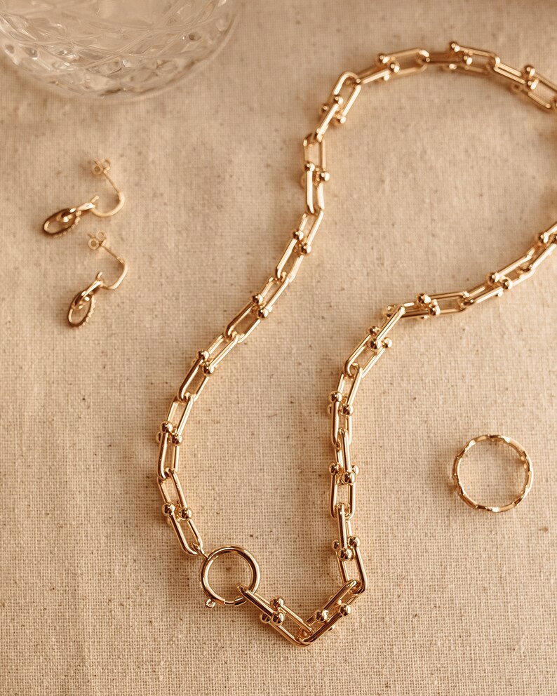 K&ouml;nnte beinhalten: Ein goldfarbenes Schmuckset mit einer klobigen Kette mit gro&szlig;em Verschluss, passenden Creolen und einem strukturierten Ring. Die Teile sind auf einer neutralen Oberfl&auml;che angeordnet und erzeugen eine zusammenh&auml;ngende und elegante &Auml;sthetik.