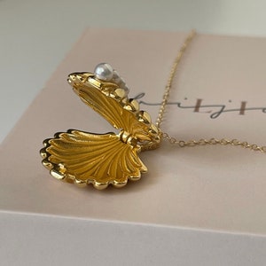Op de afbeelding: Een gouden ketting met een schelp-hanger die opengaat om een ​​klein vak te onthullen. De hanger is versierd met witte parels.