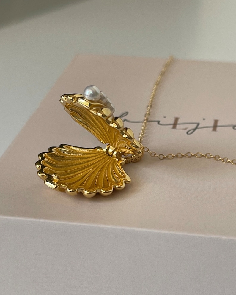 Op de afbeelding: Een gouden ketting met een schelp-hanger die opengaat om een ​​klein vak te onthullen. De hanger is versierd met witte parels.