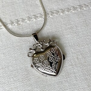 May include: A silver heart-shaped locket necklace with a delicate chain. The locket features an engraved floral design.