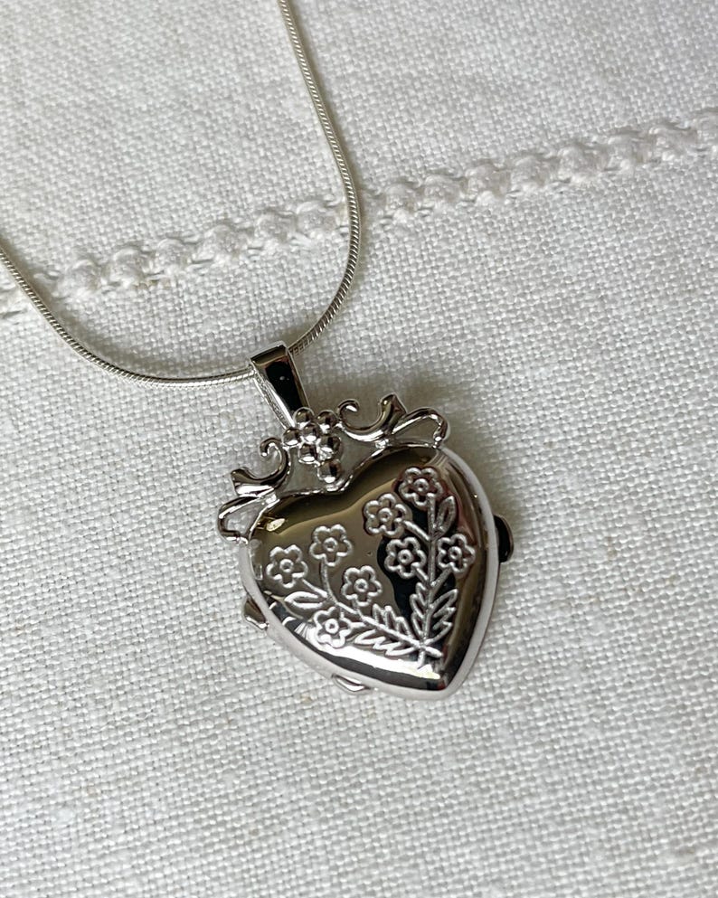 May include: A silver heart-shaped locket necklace with a delicate chain. The locket features an engraved floral design.