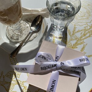 K&ouml;nnte beinhalten: Ein Caf&eacute;-Szene mit einem Glas Kaffee mit Sahne, einem Glas Wasser und einer kleinen Geschenkbox, die mit einem wei&szlig;en Band gebunden ist. Auf dem Band steht "BIJOU LIMON". Die Szene spielt sich auf einem Tisch ab.