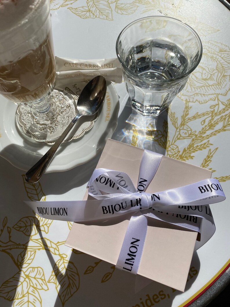 K&ouml;nnte beinhalten: Ein Caf&eacute;-Szene mit einem Glas Kaffee mit Sahne, einem Glas Wasser und einer kleinen Geschenkbox, die mit einem wei&szlig;en Band gebunden ist. Auf dem Band steht "BIJOU LIMON". Die Szene spielt sich auf einem Tisch ab.