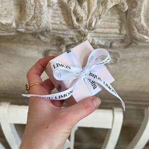 Puede incluir: Una peque&ntilde;a caja de regalo rosa claro atada con una cinta blanca con el texto "Bijou Limon" en negro. La caja se sostiene en una mano, con un anillo de oro y una pulsera de perlas visibles. El fondo es una superficie texturizada de color claro.