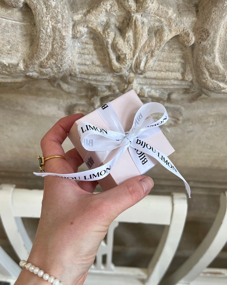 Puede incluir: Una peque&ntilde;a caja de regalo rosa claro atada con una cinta blanca con el texto "Bijou Limon" en negro. La caja se sostiene en una mano, con un anillo de oro y una pulsera de perlas visibles. El fondo es una superficie texturizada de color claro.