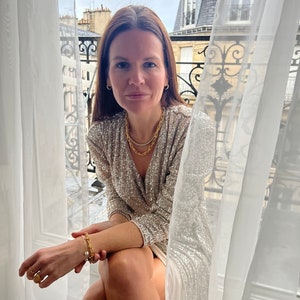 K&ouml;nnte beinhalten: Eine Frau tr&auml;gt ein goldfarbenes Pailletten-Wickelkleid und Goldschmuck, darunter eine Halskette, ein Armband und Ohrringe. Das Kleid hat einen V-Ausschnitt und lange &Auml;rmel. Die Frau sitzt in der N&auml;he eines Fensters mit wei&szlig;en Vorh&auml;ngen und Blick auf eine Stadt.