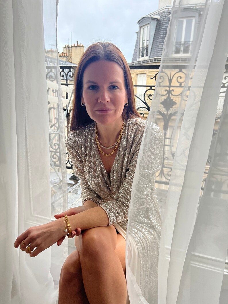 K&ouml;nnte beinhalten: Eine Frau tr&auml;gt ein goldfarbenes Pailletten-Wickelkleid und Goldschmuck, darunter eine Halskette, ein Armband und Ohrringe. Das Kleid hat einen V-Ausschnitt und lange &Auml;rmel. Die Frau sitzt in der N&auml;he eines Fensters mit wei&szlig;en Vorh&auml;ngen und Blick auf eine Stadt.