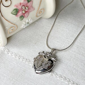 May include: A silver heart-shaped locket necklace with a delicate floral design. The locket is on a silver chain.