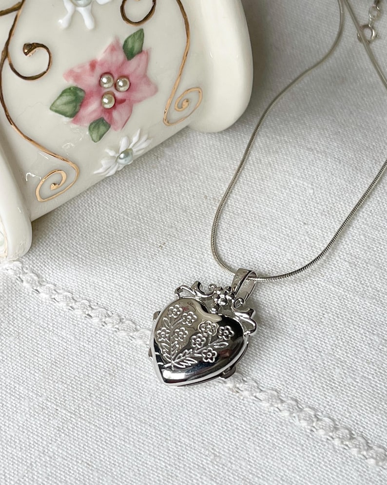 May include: A silver heart-shaped locket necklace with a delicate floral design. The locket is on a silver chain.