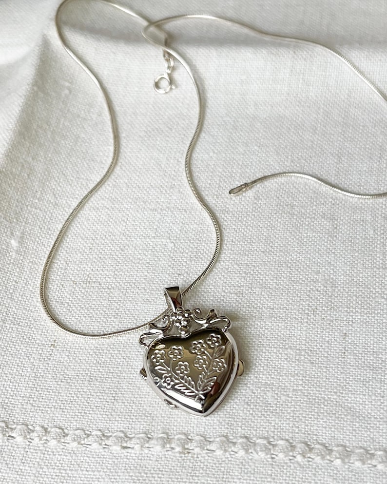 May include: A silver heart-shaped locket necklace with a delicate floral design. The chain is a snake chain style.