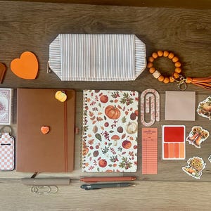 Pode incluir: Uma coleção de artigos de papelaria e acessórios, incluindo um estojo às riscas, um porta-chaves com contas cor de laranja, um caderno castanho, um caderno de espiral com tema de outono, cartas de jogar, bálsamo labial, canetas e autocolantes.