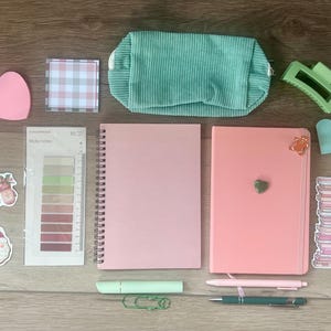 Puede incluir: Una colección de artículos de papelería que incluye un cuaderno de espiral rosa, un diario encuadernado rosa, un estuche de pana verde menta, una pinza para el pelo verde menta, un cepillo rosa, un llavero verde menta, pegatinas, notas adhesivas y bolígrafos.