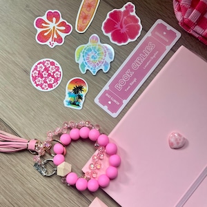 May include: A collection of pink-themed items including a light pink notebook with a heart charm, a pink beaded wristlet keychain with a tassel, and several stickers featuring a surfboard, hibiscus flowers, a sea turtle, and a ticket reading BOOK GIRLIES.