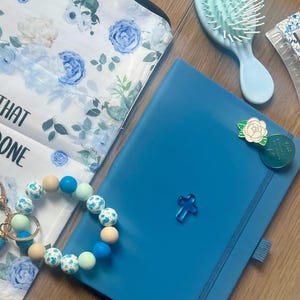 May include: A blue notebook with a small blue cross, a white rose pin, and a green circular pin. Beside it is a beaded keychain with blue, white, and tan beads, a floral patterned fabric pouch, and a light blue hairbrush with white bristles.