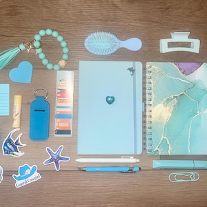 Op de afbeelding: Een verzameling lichtblauwe en talingkleurige kantoorartikelen en accessoires, waaronder een kaartspel, een kralen sleutelhanger, een haarborstel, een haarklem, een notitieboek, een spiraalplanner, een etui, stickers en diverse pennen.