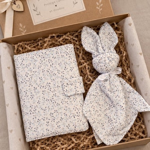 May include: A gift box containing a notebook cover and a rabbit security blanket, both featuring a blue and white floral pattern. The box lid has a cream ribbon and a label with the text Un joli coffret, Protège carnet et doudou.