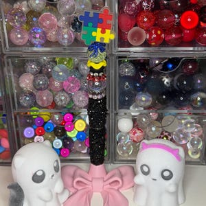 May include: A vertical pen stand featuring a pink bow base, a central column of black rhinestones and colourful beads, topped with a multi-coloured puzzle piece icon. Flanking the stand are two white ghost-like figures with large eyes.