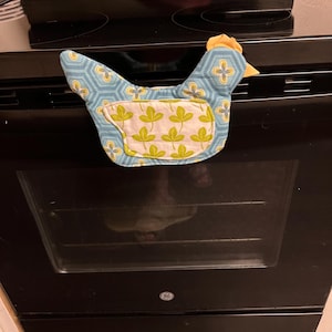 May include: A chicken-shaped fabric oven mitt featuring a light blue background with a dark blue geometric hexagon pattern and a white pocket section decorated with a repeating green leaf print. The top of the mitt has a yellow fabric comb.