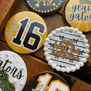 May include: A collection of six circular pin-back buttons featuring various designs, including the text "game day", "geau gator", "just here for halftime", "tors", and the number "16" with the name "Haydel" and "18" with "Haydel".