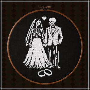 May include: A circular wooden embroidery hoop containing black fabric with a white cross-stitch design of two skeletons dressed as a bride and groom holding hands. A small white heart is stitched above them, and two rings are stitched below.
