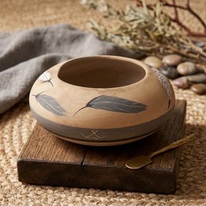 May include: A round, low-profile ceramic bowl with a matte tan finish, featuring hand-painted grey and white feather motifs around the exterior. The lower portion of the bowl is accented with a wide, dark grey horizontal band and thin line details.