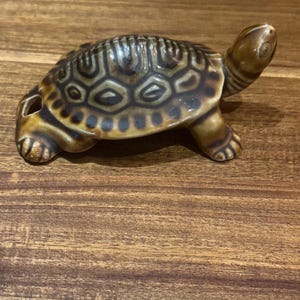 May include: A ceramic turtle figurine featuring a glossy brown glaze with darker brown, patterned markings on its shell. The turtle is depicted in a walking position with its head extended upward and a small circular opening visible on its rear shell.