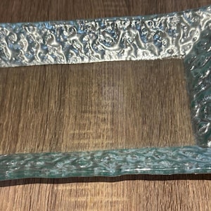 May include: A rectangular glass tray featuring a clear, light blue tint and a highly textured, uneven surface with a raised, organic pattern along the wide border. The center of the tray is smooth and transparent, showcasing a distinct relief design.