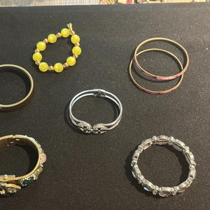 May include: A collection of six bracelets including a yellow beaded stretch bracelet, a plain gold-tone bangle, two thin copper-colored bangles, a silver-tone hinged cuff with crystal accents, a wide gold-tone buckle cuff, and a silver-tone crystal link bracelet.