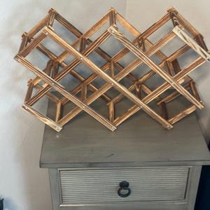 May include: A foldable wooden wine rack with a scorched finish, featuring a lattice design composed of intersecting slats connected by small metal rivets. The structure expands into a diamond-shaped grid pattern to hold multiple bottles securely.