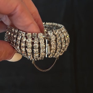 May include: A wide silver-tone cuff bracelet featuring five parallel rows of clear, faceted rhinestones. The bracelet includes a hinged metal clasp mechanism and a thin, delicate safety chain attached to the underside to secure the closure.