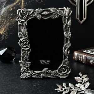 May include: A rectangular picture frame made of silver-toned metal with a raised relief design of roses, rosebuds, and leaves along the border. The center is filled with black velvet, and a small white sticker reads MADE IN TAIWAN at the bottom.