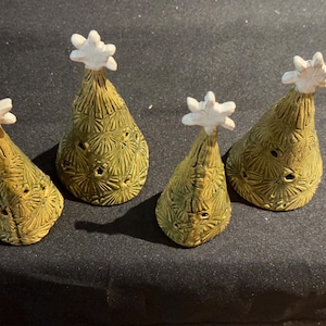 May include: Four cone-shaped ceramic ornaments in a muted olive green colour, each featuring a textured surface with small cut-out star shapes and a white, six-pointed star topper. The ornaments are arranged in a row on a dark surface.