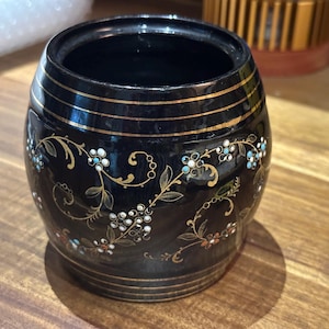 May include: A small, barrel-shaped black container with a glossy finish. The exterior is decorated with delicate gold-colored scrolling vine patterns and small, multi-colored floral accents. Three thin gold horizontal bands encircle the top and bottom.