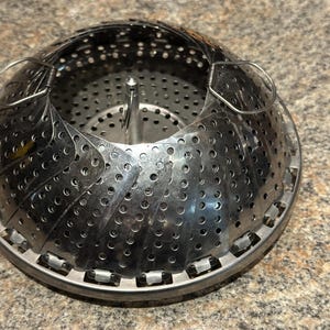 Puede incluir: Una cesta vaporera circular de acero inoxidable con pétalos metálicos perforados y superpuestos que forman una cúpula. El utensilio incluye un poste central vertical como asa y dos pequeñas anillas de alambre en el borde exterior para elevarlo.