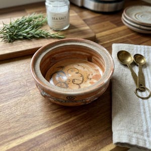 Peut inclure: Un pot à sel en céramique ronde, tourné à la main, avec une large ouverture et un vernis naturel en terre cuite. L'intérieur présente une finition lisse et brillante avec de subtils motifs tourbillonnants bruns, tandis que l'extérieur est texturé.