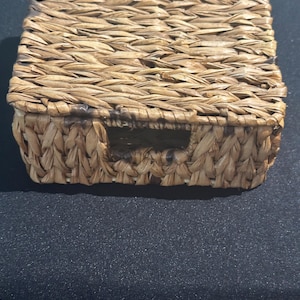 May include: A square, lidded box crafted from woven natural seagrass fibers in a light brown tone. The container features a textured, braided pattern throughout the exterior and a small rectangular cutout on the front side of the base section.