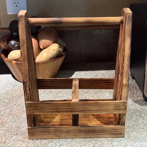 May include: A rectangular wooden carrier with a dark, scorched finish and a central handle. The structure features two open compartments separated by a vertical divider, with horizontal slats forming the sides and a solid base for stability.