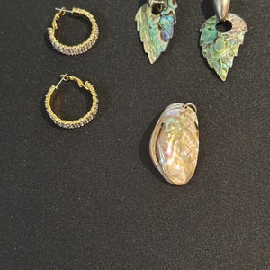 Peut inclure: Deux créoles dorées avec de petites pierres transparentes, deux boucles d'oreilles en forme de feuille avec une incrustation de nacre d'ormeau irisée verte et bleue, et un pendentif ovale avec une surface en nacre d'ormeau irisée et une bélière.