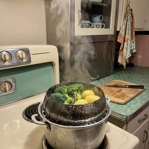 May include: A stainless steel metal steamer basket sits inside a metal pot on a stove burner. The steamer contains green broccoli florets and yellow potatoes, with visible steam rising from the pot. The stove is cream and light green.