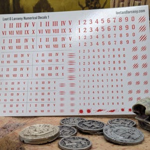 May include: A white decal sheet titled Loot & Larceny Numerical Decals 1 featuring rows of red Roman numerals from I to X and Arabic numerals from 1 to 0. The sheet includes small red slash marks and splatter designs, with the website lootandlarceny.com printed in the top right corner.
