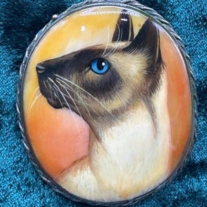 Könnte beinhalten: Eine ovale Brosche mit einem handgemalten Porträt einer Siamkatze im Profil. Die Katze hat ein dunkelbraunes Gesicht, cremefarbenes Fell und leuchtend blaue Augen. Der Hintergrund ist ein Farbverlauf in Orange und Gelb. Das Stück hat einen Silberrahmen.