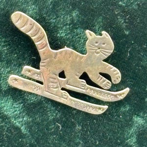 May include: A gold-toned metal brooch shaped like a cat wearing skis. The cat has engraved stripes on its body and tail, with a raised head and whiskers. The skis feature small star cutouts at the tips and textured detailing along the length.