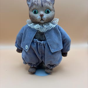May include: A cat figurine with a brown and gray striped head, green eyes, and a small brown nose. The cat wears a blue velvet jacket with a single white button, matching blue velvet bloomers, and a white lace collar around its neck.