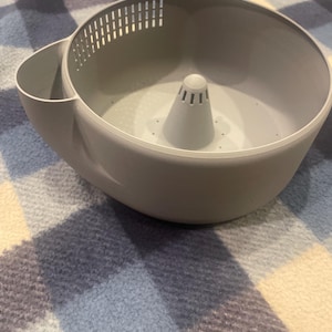 May include: A light gray plastic kitchen colander bowl featuring a central cone-shaped drainage tower, a side pouring spout, and a series of vertical drainage slots along the upper interior rim for efficient liquid removal during food preparation.